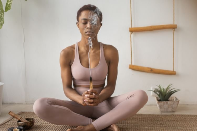 A woman meditates and burns sage as she prioritizes self-care. Self-care is one of many wedding wellness tips for brides.